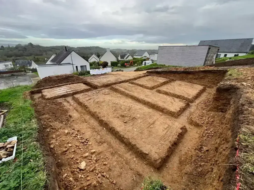 Fouilles en rigole pour semelles filantes creusées dans la terre ocre, vue sur le lotissement en contrebas, terrassement en Finistère Sud